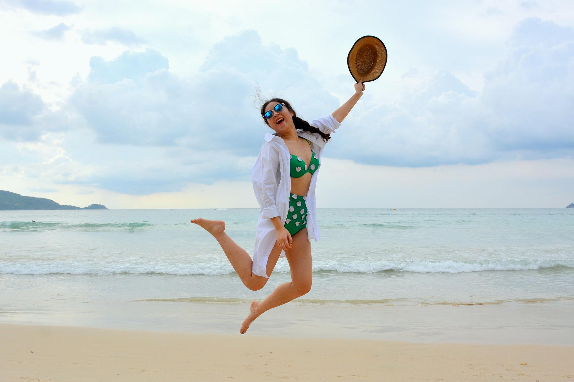 woman jumping on seashore and holding hat